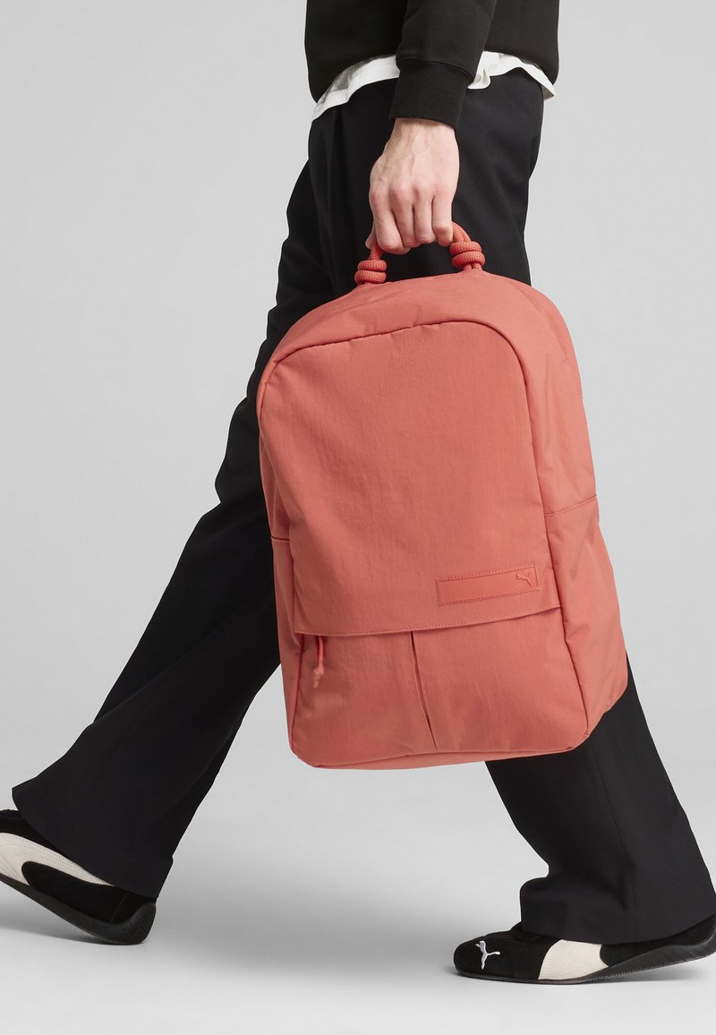 Zaino corallo con forma arrotondata, tessuto texture, maniglia superiore per il trasporto e tasca con pattina frontale. Abbinato a pantaloni neri e scarpe slip-on.