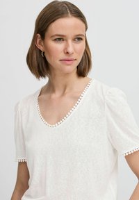 Haut blanc à manches courtes avec un décolleté en V profond, présentant une bordure décorative en forme de vagues le long du décolleté et des manches, fabriqué en tissu texturé doux.