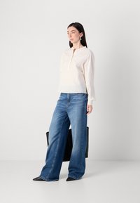 Una donna si trova in un ambiente luminoso, indossando una blusa color crema e jeans blu a gamba larga, mentre tiene una grande borsa nera.