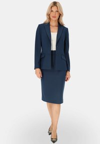 Granatowy garnitur biznesowy z dopasowaną marynarką z klapami typu notch i ołówkową spódnicą, w zestawie z białą bluzką oraz czarnymi szpilkami z złotymi akcentami.
