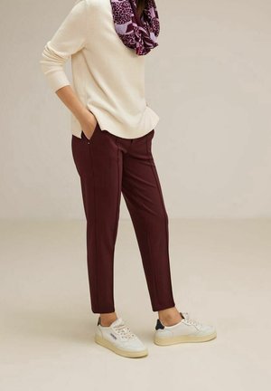 Personne portant un pull crème, une écharpe violette à motifs, un pantalon bordeaux et des baskets blanches, debout les mains dans les poches.