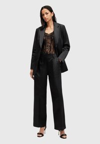 Completo in satin nero composto da un blazer strutturato e pantaloni a gamba larga. Body in pizzo sotto con motivi intricati. Sandali neri con tacco.