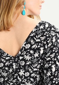 Robe noire à fleurs avec un décolleté en V ornée de motifs botaniques blancs. Des boucles d'oreilles en turquoise en forme de goutte d'eau complètent la tenue.