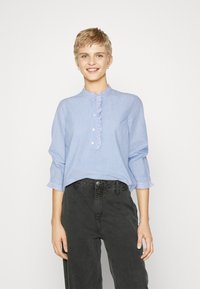 Lyseblå,stripete langermet skjorte med volangkrage og knappelukking; har samlete mansjetter. Kombinert med svarte høyde i livet jeans.