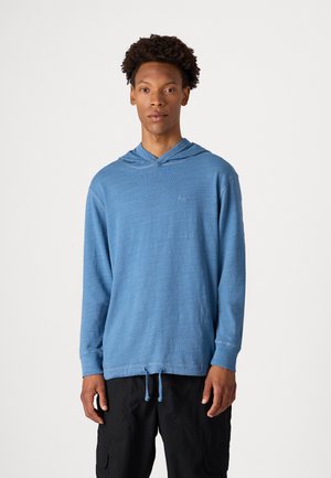 Lichtblauwe hoodie met lange mouwen en een koord, van gestructureerde stof met een subtiele logodetail op de borst. In combinatie met donkere, loszittende broeken.