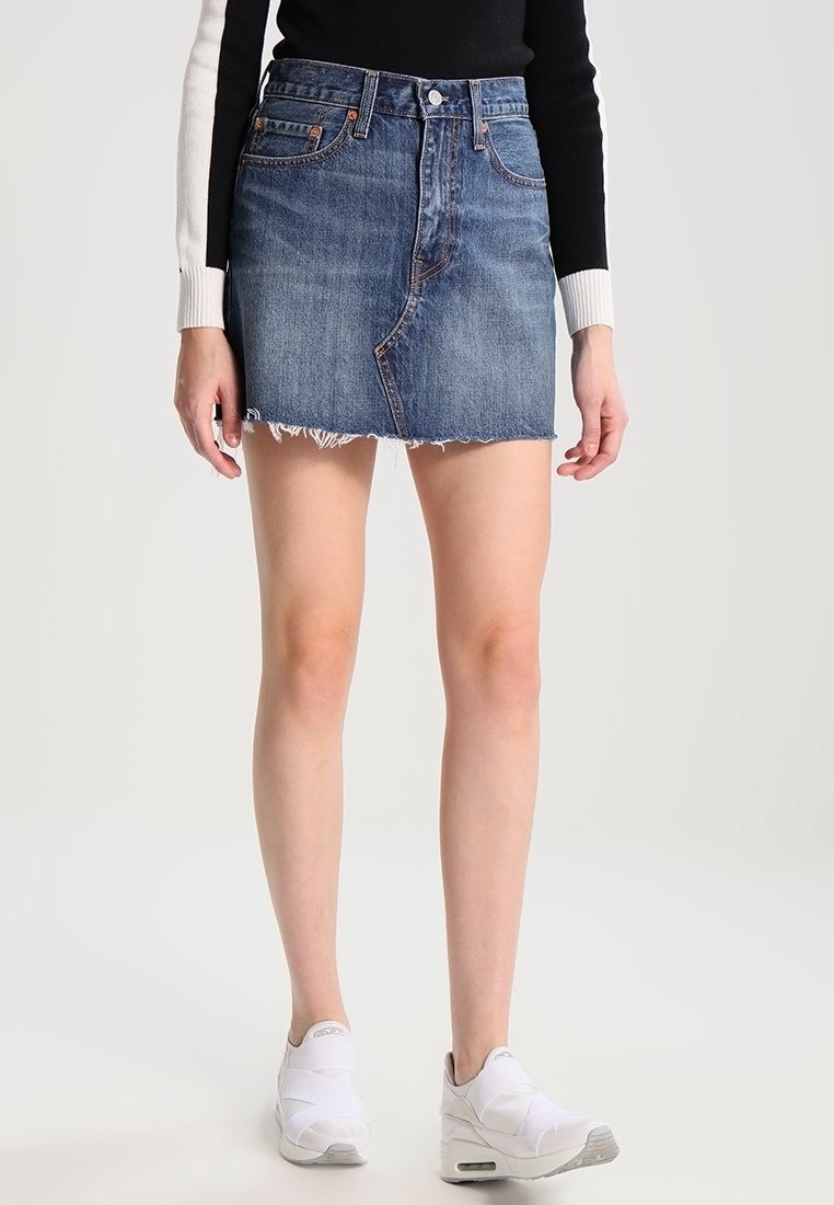 Femme portant une mini-jupe en jean bleu avec un ourlet brut, un top à manches longues noir et blanc et des baskets blanches, se tenant sur un fond clair.