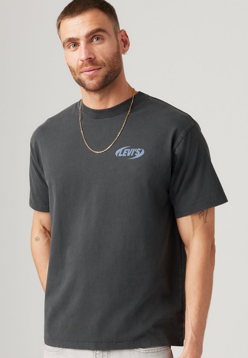 Mann mit kurzen Haaren und Bart, der ein schwarzes Levi's T-Shirt, eine goldene Kette und Ohrringe trägt, steht vor einem schlichten hellen Hintergrund.
