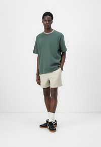 Dark green short-sleeve T-shirt with a contrasting white and green ribbed neckline, paired with beige shorts and black sneakers.