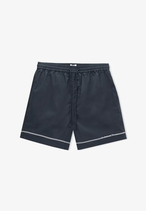 Ljetne tamnoplave hlačice s bijelim izvezenim natpisom i detaljem od tri linije uz rub na nozi osobe na jednobojnoj pozadini.