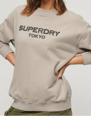 Béžová mikina s černým nápisem „SUPERDRY TOKYO“, kterou nosí osoba se založenýma rukama v bok a olivově zelenými kalhotami.