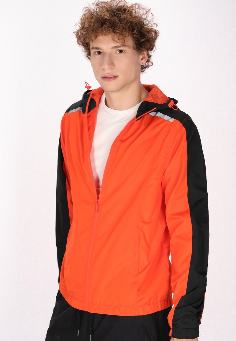 Orange og sort lynlåsjakke med hætte, reflekterende detaljer, letvægtsmateriale og tætsiddende design.