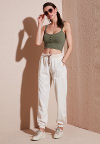 Groene geribbelde crop top met drie knopen, gecombineerd met witte elastische broek met zakken en een trekkoord. Grijze sneakers maken de look af.