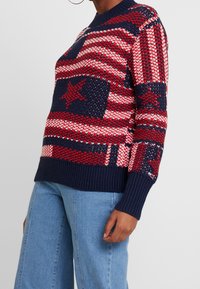 Persoon in een gebreide trui in rood, wit en marineblauw met sterren- en streepjespatronen, gecombineerd met blauwe jeans die halverwege de torso zijn afgeknipt.