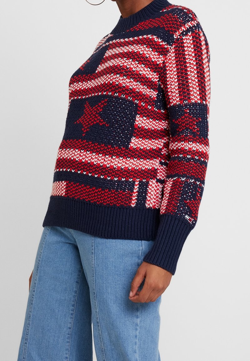 Persoon in een gebreide trui in rood, wit en marineblauw met sterren- en streepjespatronen, gecombineerd met blauwe jeans die halverwege de torso zijn afgeknipt.