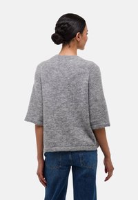 Femme avec des cheveux bruns en chignon, portant un pull en maille gris à manches courtes et un jean bleu, de dos sur un fond blanc uni.