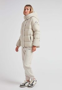 Puffjakke i lys beige med høj krave og store paneler. Sammen med matchende beige joggers og sort-hvide sneakers.