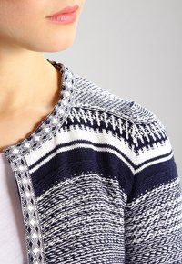 Närbild av en person som bär en marinblå och vit mönstrad stickad cardigan över en vit topp, som visar nacke och axelområde.