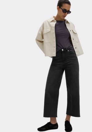 Femme portant des lunettes de soleil noires, une veste beige, un t-shirt gris foncé, un jean large noir et des baskets noires, posant devant un fond clair uni.
