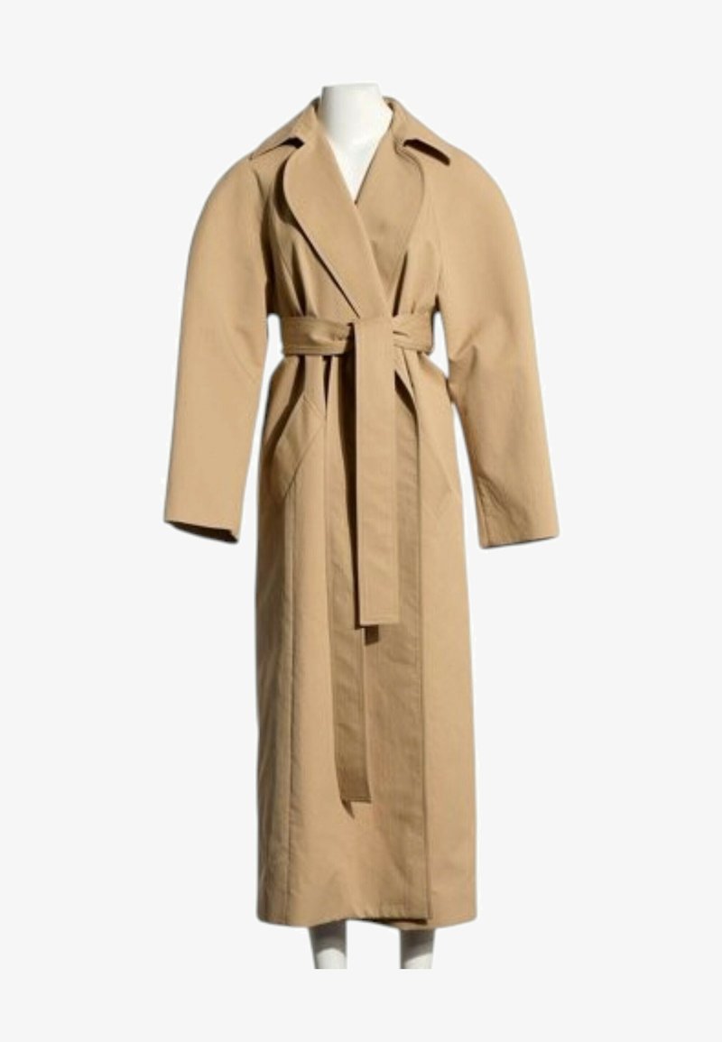 Long trench-coat beige avec larges revers, grandes manches, poches latérales et ceinture assortie à la taille sur un mannequin blanc.