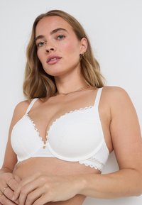 Reggiseno bianco imbottito con dettagli in pizzo a piccole onde e spalline regolabili. Presenta una texture liscia e un piccolo bottone decorativo al centro.