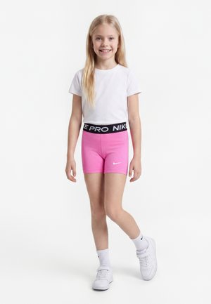 Uśmiechnięta młoda dziewczyna stoi ubrana w białą koszulkę, różowe spodenki Nike Pro, białe skarpetki i białe tenisówki na jednolitym białym tle.