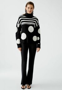 Gestreifter Pullover in Schwarz-Weiß mit Polka-Dots und hohem Kragen, aus strukturiertem Strick. Kombiniert mit schwarzen Hosen und flachen Sandalen.