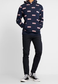 Sudadera navy con el logotipo "SCOTCH" en blanco y rojo repetido, combinada con pantalones negros ajustados y zapatillas de caña alta negras.