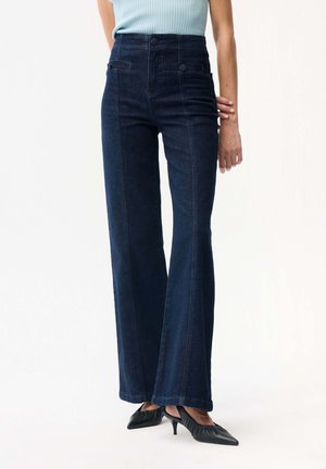 Kobieta ubrana w ciemnoniebieskie dzwony z wysokim stanem, połączone z czarnymi butami na obcasie w szpic oraz jasnoniebieską bezrękawnik.