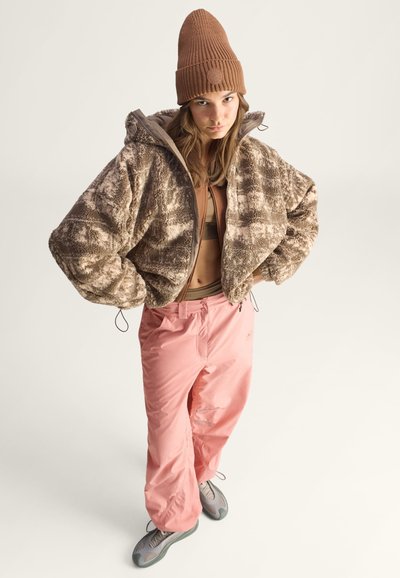 Bruine, bontachtige, cropped jas met een capuchon. Tan basislaag top. Roze baggy broek. Model draagt een bruine geribbelde beanie en grijze sneakers.