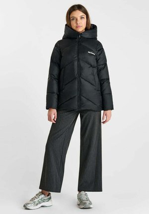 Donna in piedi che indossa una giacca imbottita nera con cappuccio e cerniera, pantaloni ampi a righe grigie e scarpe sportive argentate.