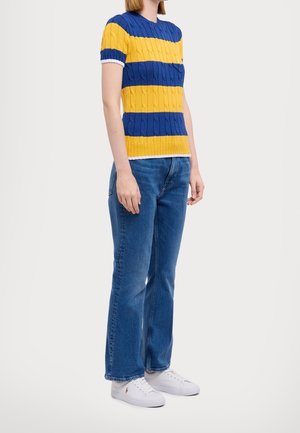 Femme portant un pull tricoté à manches courtes avec des rayures bleues et jaunes, un jean bleu et des baskets blanches, debout devant un fond uni.