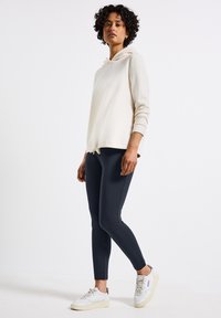 Krämfärgad huvtröja med dragsko, kombinerad med mörkblå leggings och vita sneakers med texturerade detaljer. Avslappnad sportoutfit.