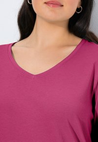 T-shirt z dekoltem w serek w kolorze fuksji, wykonany z miękkiej tkaniny bawełnianej. Wyróżnia się gładką fakturą i prostym wzornictwem, z przeszyciami wokół dekoltu.