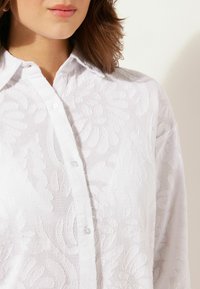 Camicia bianca con pulsanti, caratterizzata da un motivo floreale testurizzato, con colletto classico e bottoni trasparenti, esposta su uno sfondo neutro.