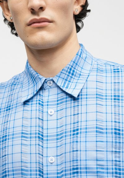 Κοντινό πλάνο ενός ατόμου που φοράει πουκάμισο με ανοιχτό μπλε καρό σχέδιο και ασημένια σκουλαρίκια καρφί, ουδέτερη έκφραση, απλό ανοιχτόχρωμο φόντο.