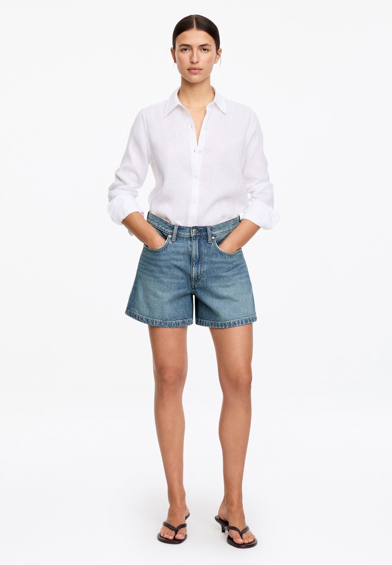 Femme debout les mains dans les poches, portant une chemise blanche à boutons, un short en jean bleu et des sandales à talons noires sur fond blanc.