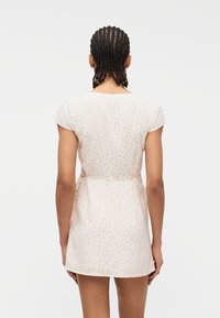 Άτομο με πλεξούδες, φοράει κοντό, ανοιχτόχρωμο φόρεμα με μικρό λουλουδάτο σχέδιο, στέκεται μπροστά σε απλό λευκό φόντο.