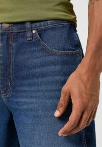 Dżinsy w ciemnoniebieskim kolorze z jasnym przeszyciem, o dopasowanym kroju, pięcioma kieszeniami i metalowymi elementami przy pasie oraz kieszeni.