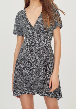 Femme portant une robe portefeuille noire à manches courtes avec un motif floral blanc et un décolleté en V, debout devant un fond uni.