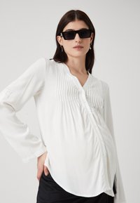 Blusa bianca a maniche lunghe con chiusura a bottoni, caratterizzata da dettagli plissettati e un orlo arrotondato. Tessuto leggero con una superficie liscia.