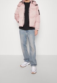 Ljusrosa pufferjacka med huva, svarta detaljer på axlarna, dragkedja fram, matchad med en svart skjorta, blå jeans och vita sneakers.