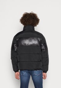 Chaqueta de plumas negra con un panel superior brillante, cuello alto y puños acanalados. Presenta un diseño acolchado y un ajuste relajado.