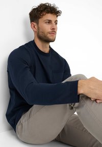 Marineblaues Langarmshirt mit Rundhalsausschnitt und Brusttasche, kombiniert mit hellbeigen Hosen. Der Stoff wirkt glatt und hat eine weiche Textur.