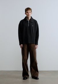Pull-over charbon à col zippé, coupe décontractée, associé à un pantalon marron ample. Tissu lisse, design minimaliste, détail de logo subtil sur le pull-over.