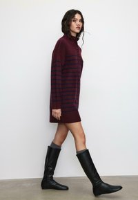 Haljina od pletiva s bordo prugama, visokim ovratnikom i detaljem s gumbima. Dugi rukavi i rebrasti rub, upareni s crnim čizmama do koljena.