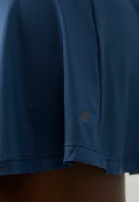 Tumši zils sporta svārku modelis, izgatavots no gludas, vieglas auduma. Tam ir līgans apakšmalas dizains un neliels tonāls logo tuvu apakšmalai.