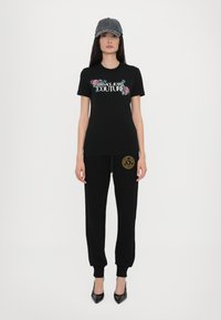 Črna bombažna majica s cvetličnim grafikonom in napisom "Versace Jeans Couture", v kombinaciji z črnimi trenirkami z zlatim logotipom in črnimi petkami.