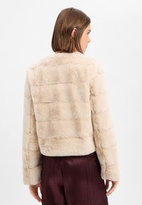 Hellbeige Kunstpelzjacke mit gestreifter Textur, kurzem Schnitt, rundem Ausschnitt und langen Ärmeln, kombiniert mit dunklen, plissierten Hosen.