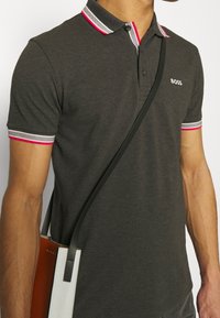 Polo gris foncé avec col contrastant à rayures blanches et roses, patte à deux boutons, porté avec un sac bandoulière.