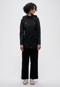 Svart vanntett jakke med glidelåslommer, justerbar hette og snor i midjen. Kombinert med svarte bukser med vide ben og hvite sneakers.